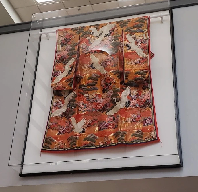 Orange kimono with white cranes and floral patterns displayed inside a wall-mounted glass case.